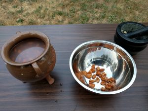 Brown table with brown pipkin on left holding almonds in water, metal bowl in center with almond peels, and black mortar and pestle on right with ground almonds