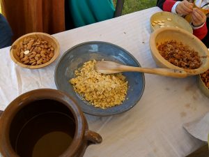 On the left is a brown pipkin with water, center is a blue bowl with ground almonds and wooden spoon. In front and to the left is a wooden bowl containing raw almonds. On the far right are two bowls, one containing almond peels and one, barely visible, containing soaked almonds. The garb of a small child can be seen.