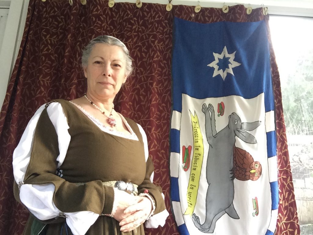 White woman, the author, standing in garb with a banner to the right, depicting a gray rabbit running with an owl riding its back, 3 red shuttles, a motto under the rabbit, and the Tir Righ 8 sided white star on a blue background
