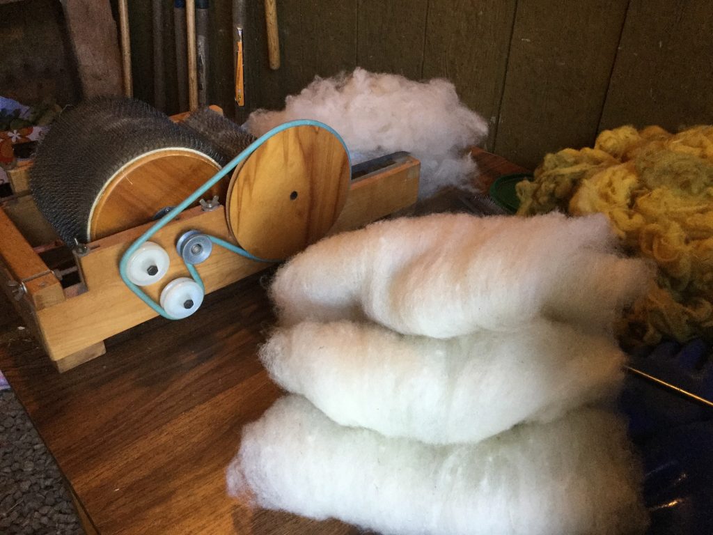 a box with two rollers with metal combers, like velcro. Wool is being fed into carder. three batts of carded fleece are in the foreground