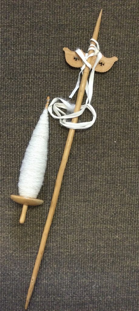 a wooden spindle with a full load of spun white wool. A long pole with a hand carved perpendicular piece of wood is near the to A ribbon is draped around the top of the distaff.