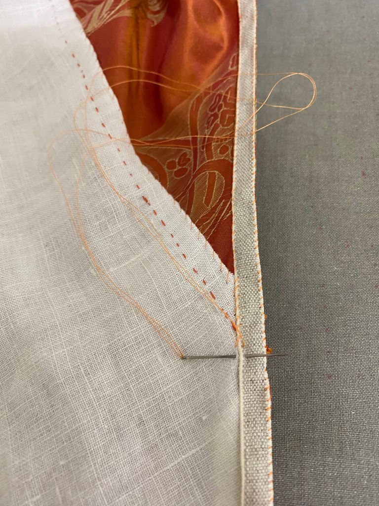 A beige linen tape is being sewn to the edge of a garment made of orange silk and white linen